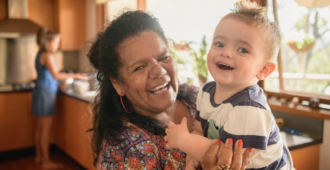 A senior woman holding her baby grandson and smiling in a kitchen.