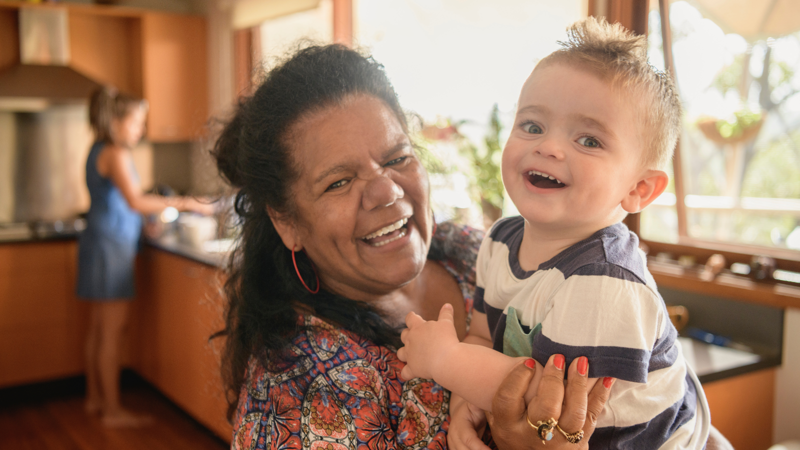 A senior woman holding her baby grandson and smiling in a kitchen.